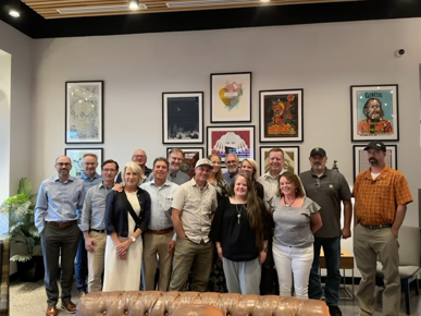A large group of people posing together in front of a wall full of picture frames.